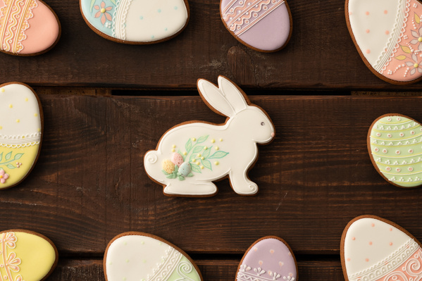 Easter glazed gingerbread cookies of different colors in the shape of an egg scattered around a rabbit-shaped gingerbread on a dark wood table