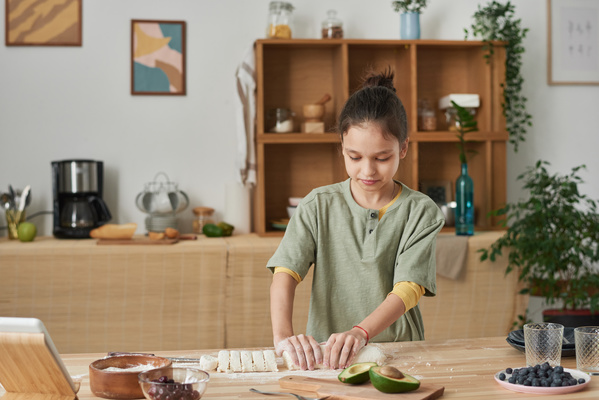 A girl with her hair in a bun shaping out a roll from dough at the kitchen table