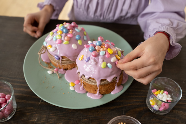 Easter cakes in pink glaze on a green plate are decorated with bright sweets by a girl in a pink blouse