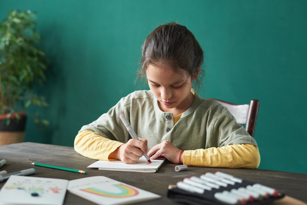 A girl with tidied up hair painting pictures on a homemade card for Fathers Day with a red felt-tip pen while sitting at a table