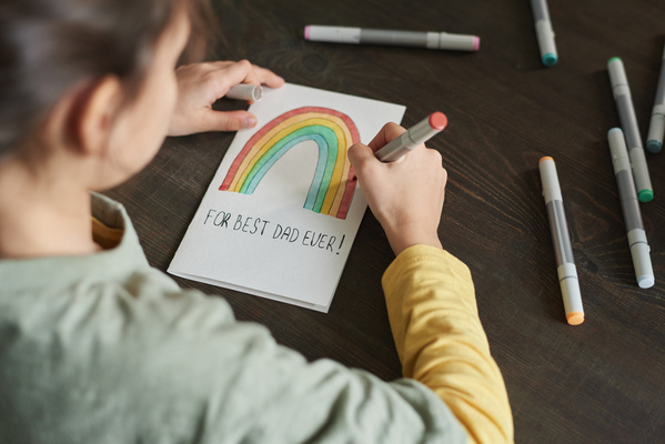 A girl with her hair tidied up coloring a rainbow on a handmade Fathers Day card with a red felt-tip pen