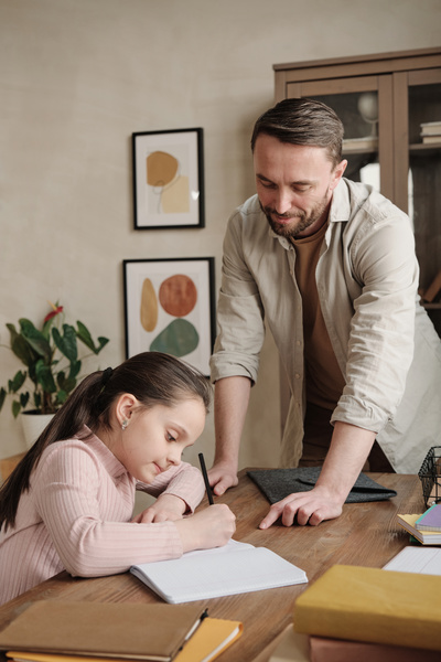 A man dressed in a beige shirt tells his daughter with her hair in a ponytail sitting at her desk how to complete a written task in a notebook