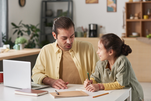 A dad in a yellow shirt communicating with his daughter holding a pencil and a notebook while sitting at a table with a laptop