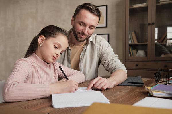 A girl with her hair in a ponytail does her homework in writing with the help of her father sitting next to her