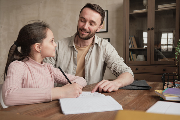 A girl with her hair in a ponytail doing her homework in writing with the help of her dad wearing a light shirt