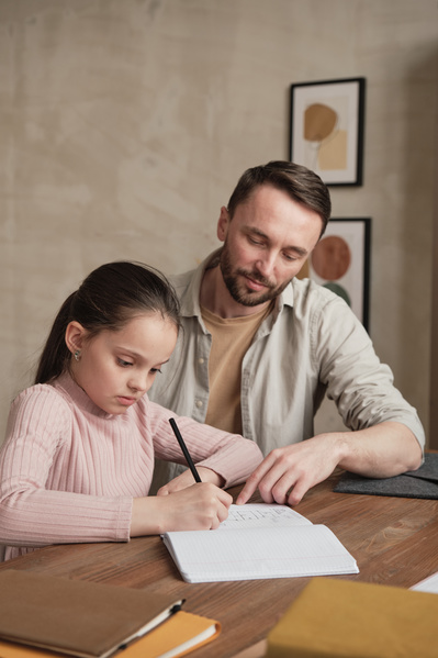 Dad in a light shirt prompts his daughter doing homework while sitting at a desk together