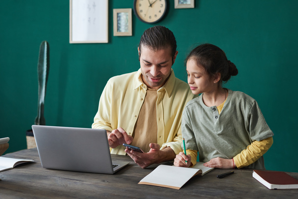 A man dressed in a yellow shirt holding a phone helps his daughter to do homework