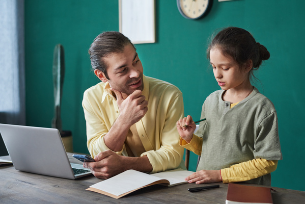 A man dressed in a yellow shirt explaining homework task to his daughter with a notebook and pencil sitting at a desk together