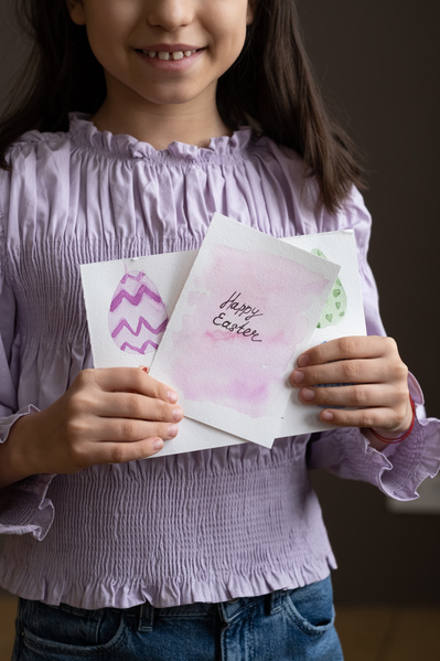 A girl with loose hair dressed in a pink blouse holding handmade Easter cards with a bright illustration and greeting