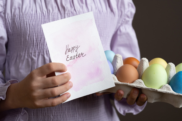 Carton with Easter eggs painted in different colors and a postcard with a wish in the hands of a girl in a pink blouse