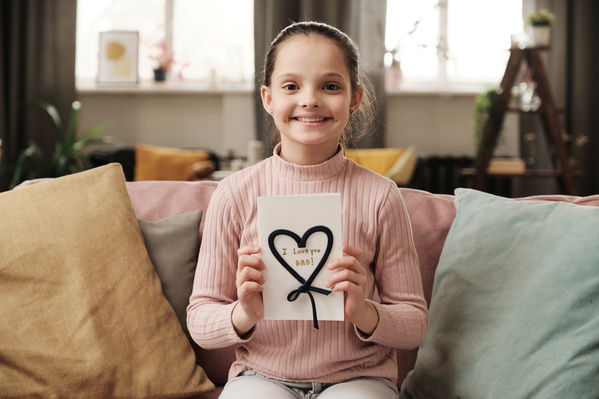 A smiling broadly girl sitting on the sofa and holding a homemade postcard for Fathers Day with the inscription and decoration in the form of a heart made of dark lace