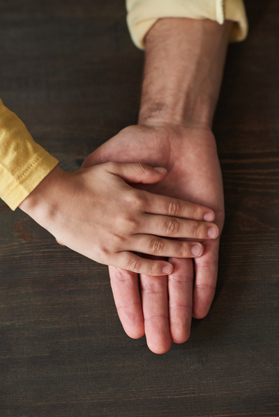 The hand of a child in a yellow longsleeve is placed on the hand of an adult man on a dark wood surface