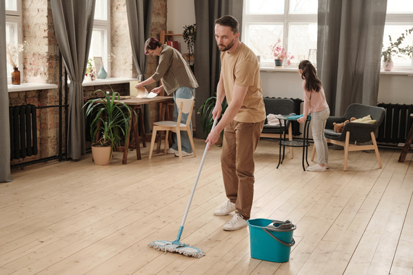 A man mopping the wooden floor next to a blue bucket and his family cleaning the room
