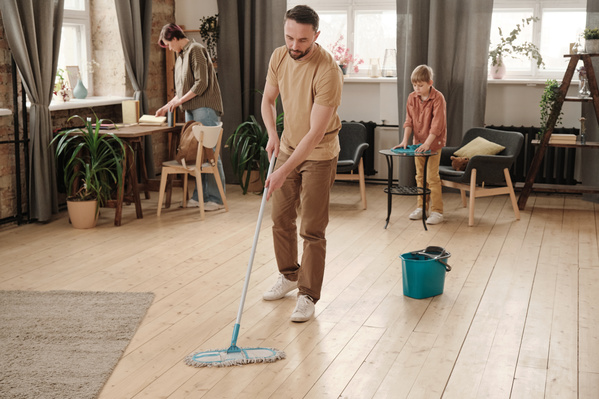 The husband in light colored clothes cleaning the wooden floor with a mop while his wife and son tidying up the room