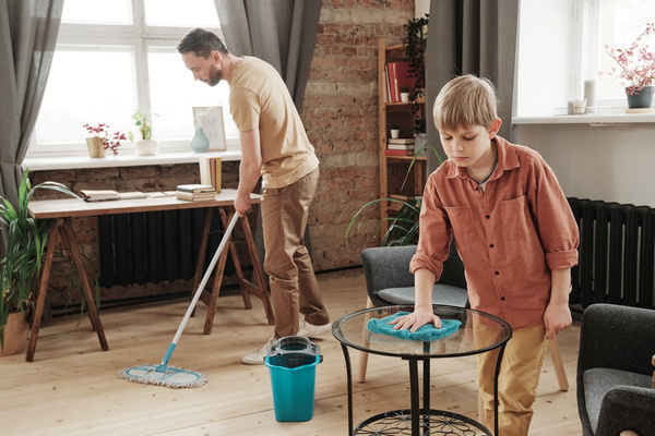 A boy with blond hair dusting the coffee table and his father mopping the wooden floor