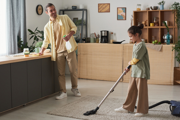 A father wiping a kitchen set looks at his daughter with her hair in a bun vacuuming a light carpet