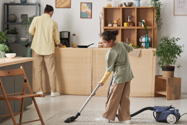 A girl with her hair in a bun vacuuming a light carpet and her father wiping the kitchen set in the kitchen
