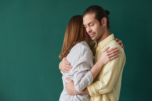 A husband in a yellow shirt hugging gently his wife in a striped blouse against an emerald-colored background