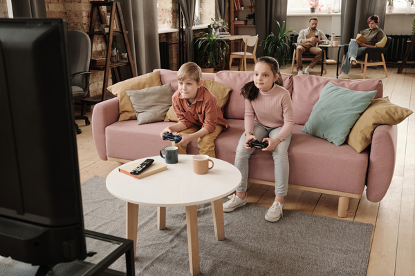 A boy in a brick-colored shirt and his sister with her hair in in a ponytail playing console with black game controllers while sitting on a pink sofa