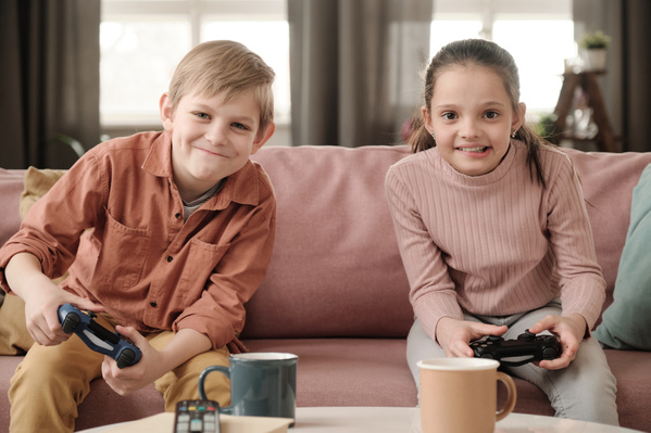 A brother in a brick-colored shirt sitting on a pink sofa together with his sister with her hair tidied up and playing console competitively with dark gamepads