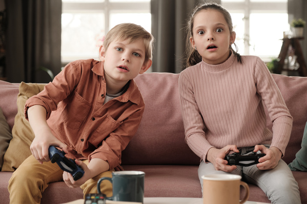 A brother in a brick-colored shirt sitting on a pink sofa together with his sister with her hair tidied up and emotionally playing video games with black game controllers