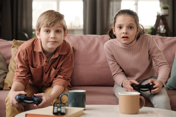 A boy in a brick-colored shirt sitting on a pink sofa and his sister with tidied up hair playing video games with emotional face expressions