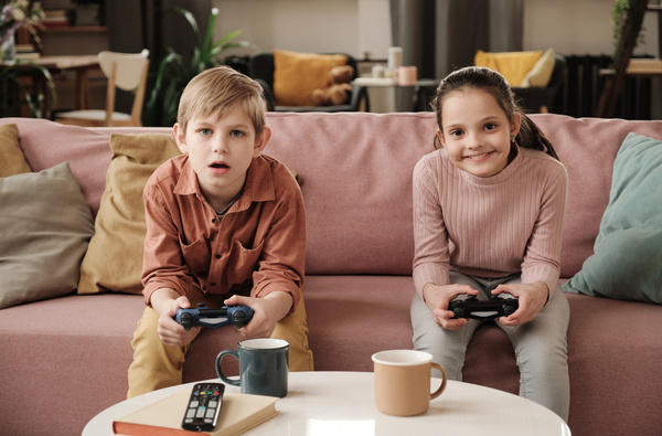 A boy in a brick-colored shirt sitting on a pink sofa loses to his sister with tidied up hair a video game