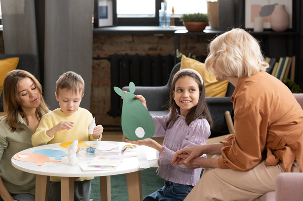 A girl in a pink blouse showing a handmade Easter card in the form of a rabbit to her grandmother while mom and son glue the application