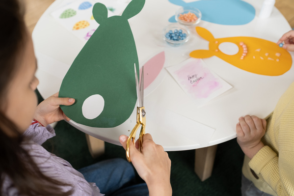 A girl cutting out green Easter cards with scissors with a golden handle sitting at a white round table