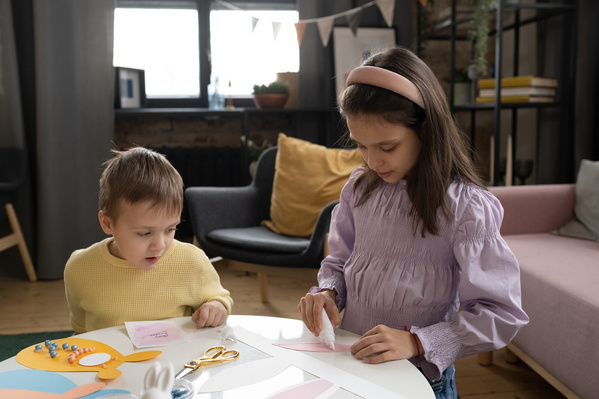 A girl in a pink blouse with her younger brother in a yellow sweater at a round white table gluing applications on Easter cards
