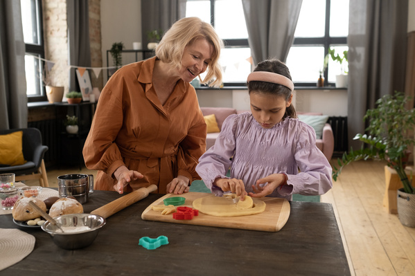 A girl in a pink blouse and her blonde-haired grandma cutting cookies for Easter out of dough