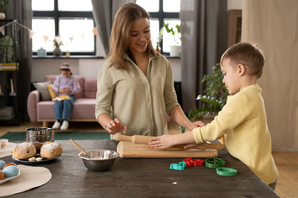 A mother in a light shirt helps her little son in a yellow sweater rolling out the dough for the Easter cookies with a wooden rolling pin