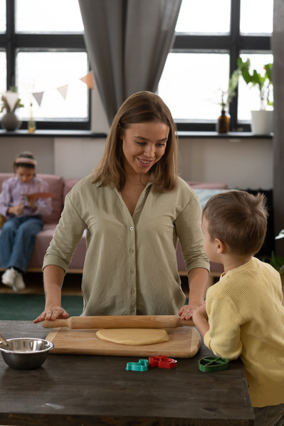 A woman in a light shirt with a young son in a yellow sweater in the kitchen rolling out the dough for Easter gingerbread with a wooden rolling pin
