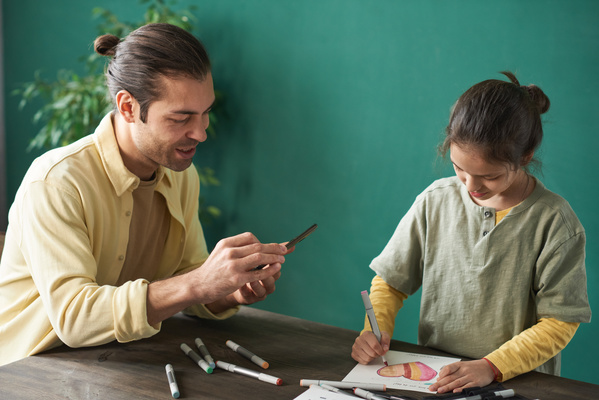 A man in a yellow shirt takes a photo on his phone of his daughter drawing illustrations on a homemade Fathers Day card