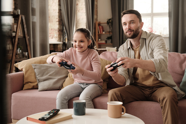A girl with tidied up hair enthusiastically plays a video game console with her father sitting on a pink sofa