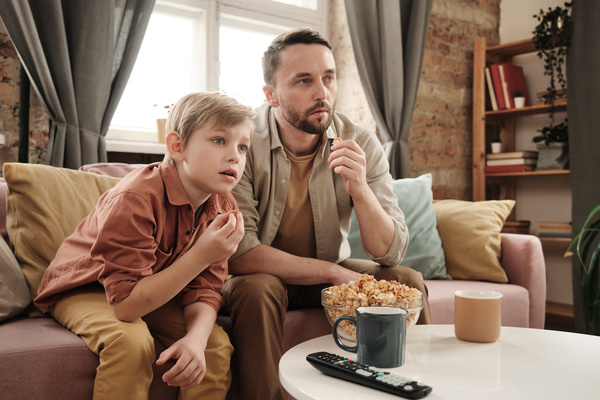 A boy with blond hair and his father eating snacks from a glass bowl watching television on a pink sofa