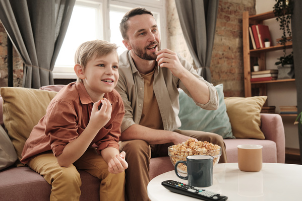 A boy with blond hair and his father eating snacks from a glass bowl enthusiastically watching a TV program on a pink sofa