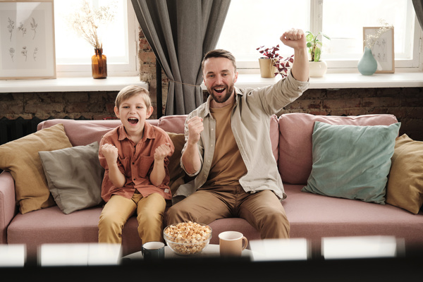 A son with blond hair and a father are happy about a goal watching football on TV sitting on a pink sofa with snacks on the table