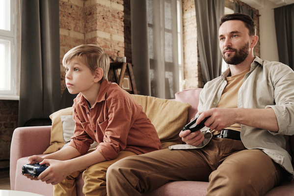 A man in a beige shirt sitting on the couch playing a video game with his blond son in a brick-colored shirt