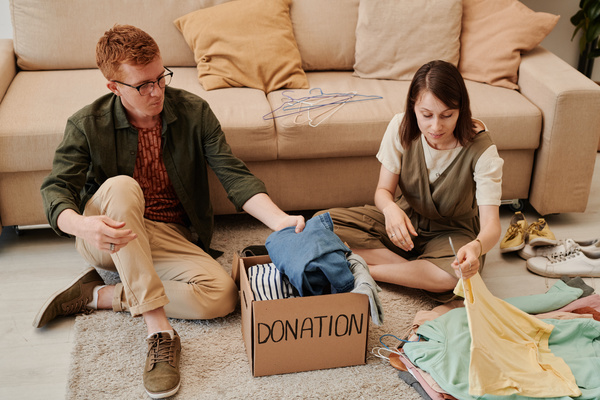A man with red hair and a woman with a square sitting on a light carpet putting clothes in a cardboard donation box