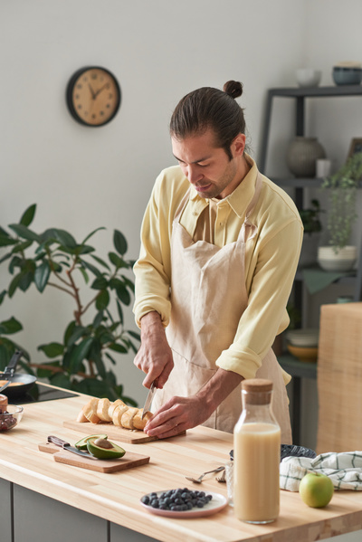 A father in a beige apron slicing a loaf of white bread on the cooking table with groceries