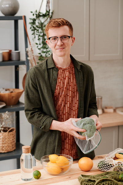 A red-haired man with glasses wearing a green shirt over a red T-shirt standing at the kitchen table with grocery purchases holding broccoli in a reusable vegetable bag