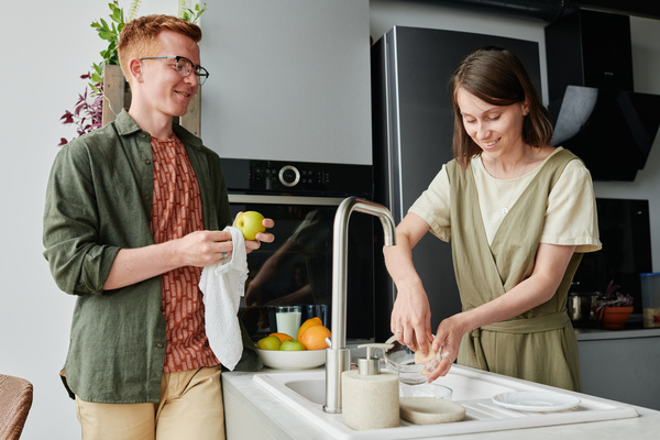 A red-haired man in a green shirt over a red T-shirt wiping an apple with a white towel and a woman with short dark hair washing dishes