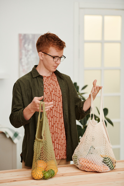 A red-haired man in glasses and a green shirt over a red T-shirt holding green and white cotton shopping bags with groceries and standing at the kitchen table