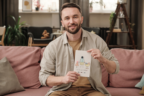 A man with a beard dressed in a light shirt sitting on a pink sofa holds a Fathers Day card with a pattern and applique in his hands
