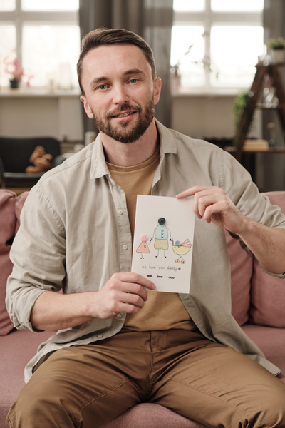 A man with a beard dressed in a light shirt sitting on a pink sofa with a postcard dedicated to Fathers Day with a pattern and applique