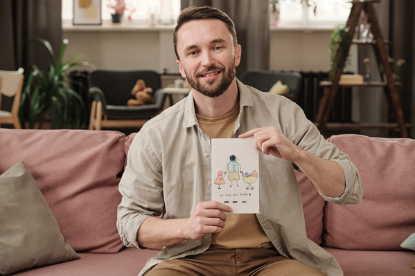 A man in a light shirt smiling sits on the sofa with a homemade postcard in honor of Fathers holiday with an illustration and an application