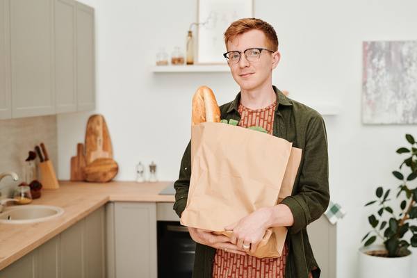 A man with red hair wearing glasses and a green shirt holds a paper bag of grocery purchases standing in a bright kitchen