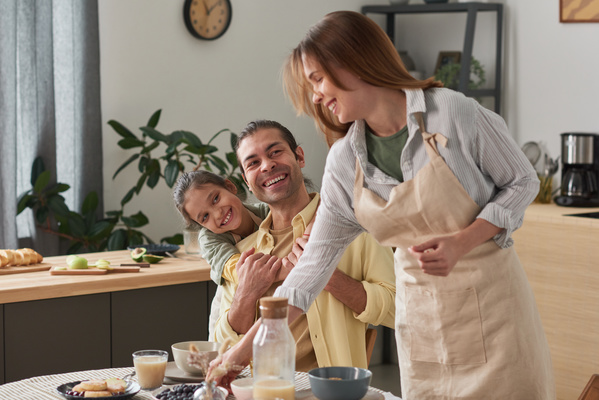 A girl with tidied up hair hugs her father sitting at the dining table and smiling at his wife wearing an apron serving the table