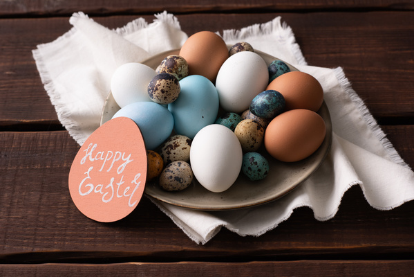 Easter chicken and quail eggs of different colors on a light plate with a greeting card on a white cloth napkin